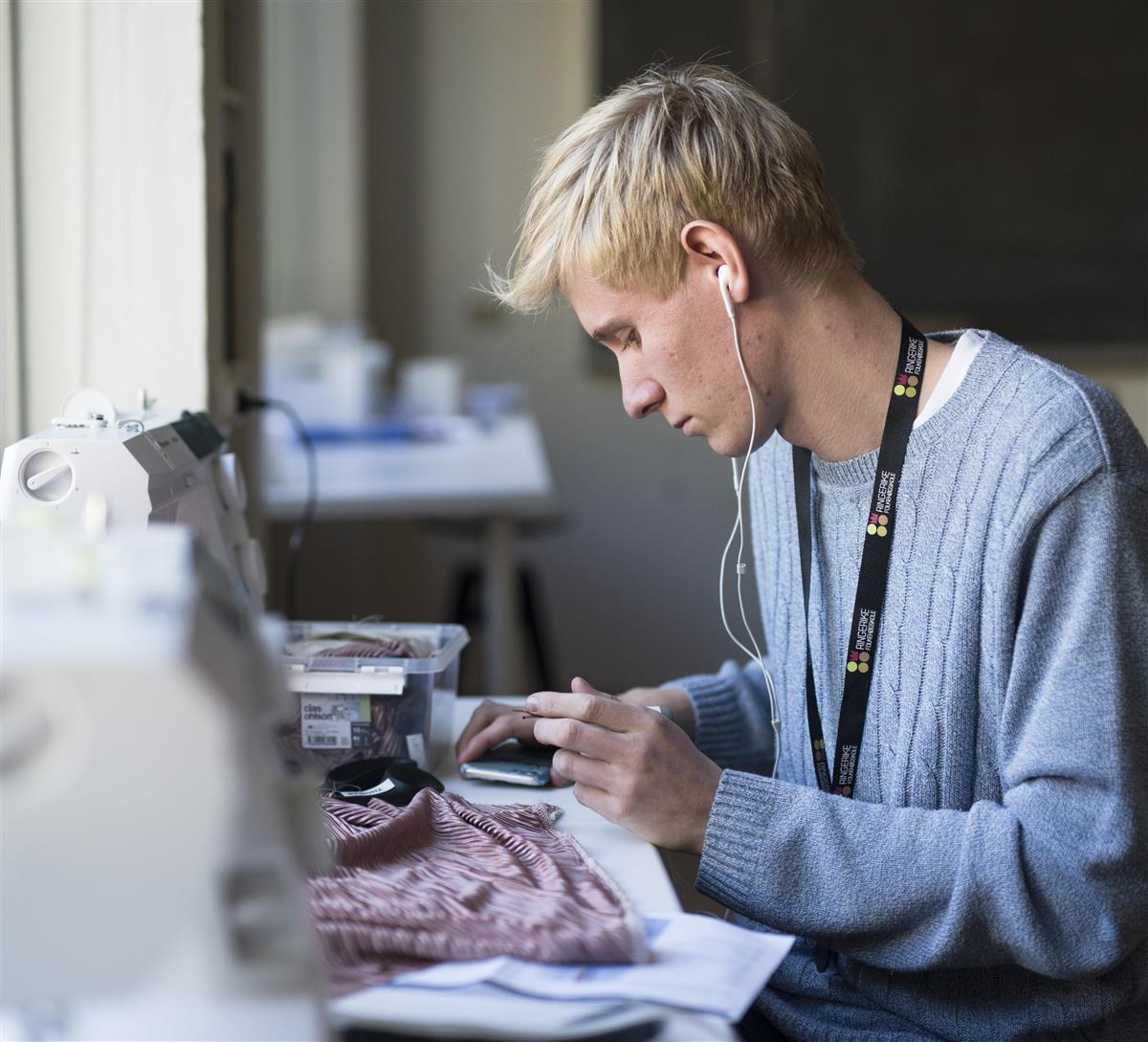 En elev sitter foran en symaskin og jobber med et syprosjekt mens han hører på musikk på øret.  - Klikk for stort bilde