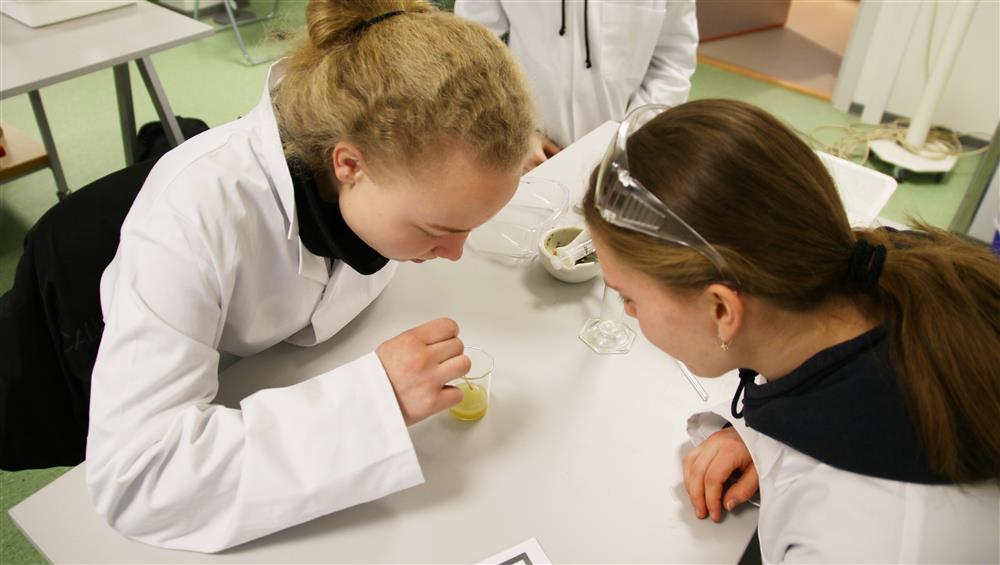 To kvinnelige elever ikledd laboratoriefrakk. Den ene rører med en pinne i et plastglass. - Klikk for stort bilde