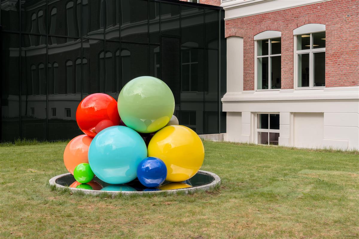 Bildet viser en skulptur bestående av kuler i ulike størrelser og farver som er plassert på en reflekterende, sirkelrund plate. Verket står midt på en gressplen, midt mellom et eldre og et helt moderne glassbygg. - Klikk for stort bilde