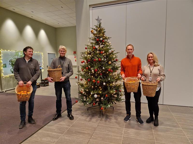 Fire ansatte ved siden av et juletre. I hendene holder de hver sin kurv med klementiner - Klikk for stort bilde